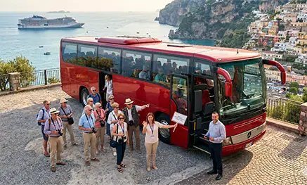 A group of cruise ship passengers on a shore excursion boarding a red Mercedes private coach bus on a scenic Amalfi Coast overlook with a cruise ship in the background