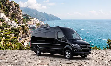 A premium executive black Mercedes-Benz Sprinter minibus parked on a stone panoramic terrace overlooking the Amalfi Coast sea and Ravello cliffs