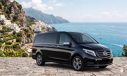 A premium luxury black Mercedes-Benz V-Class minivan parked on a stone panoramic terrace overlooking the Amalfi Coast sea and Positano cliffs