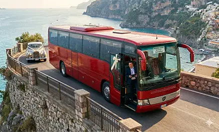 A luxury red Mercedes coach bus coordinating wedding guest transportation alongside a vintage bridal car on a scenic Amalfi Coast cliffside road